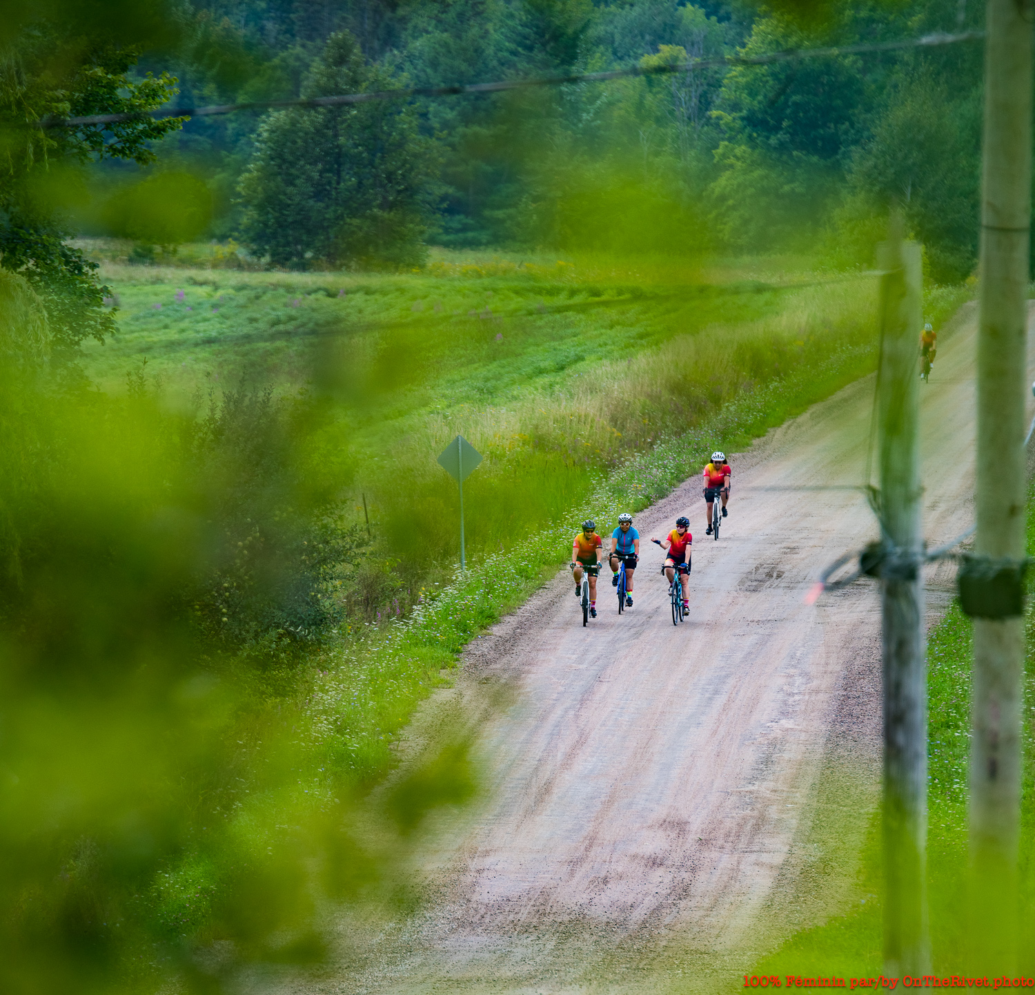 A group of women in OWCC jerseys ride down a gravel road with lush greenery on the sides.